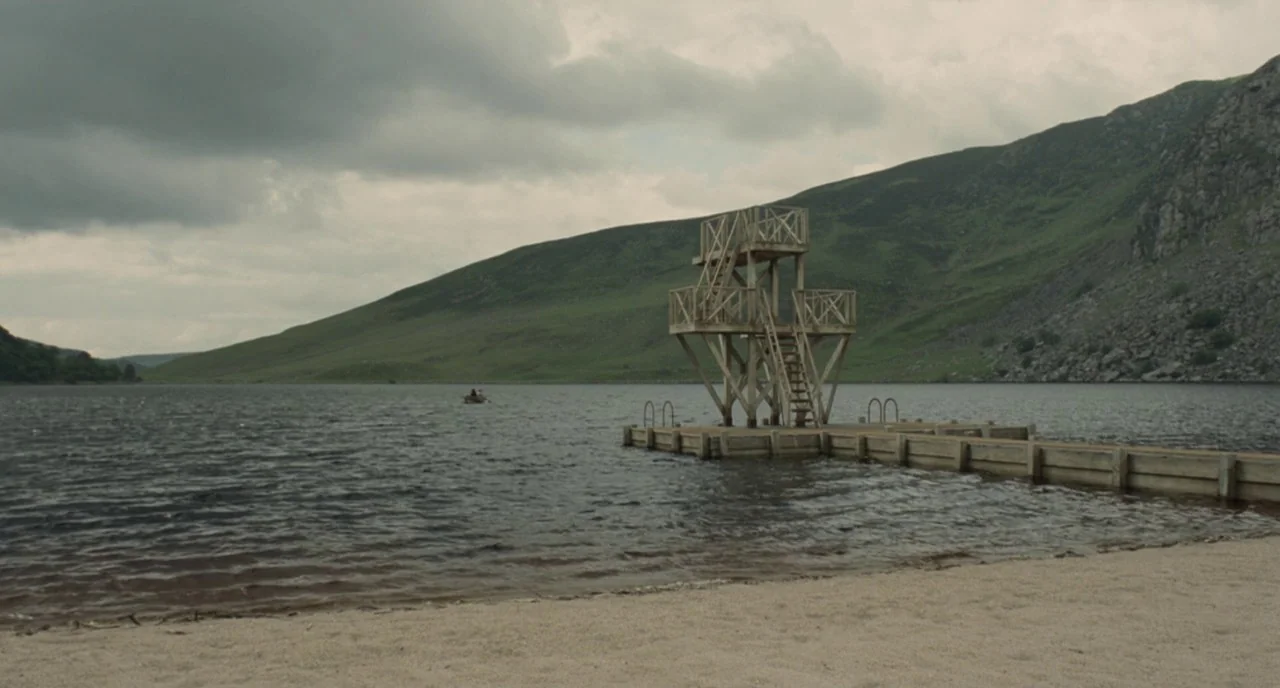 Cracks (2009) movie still: wide — A wooden diving tower sits at the end of a pier on a lake. Rolling green hills rise in the background under a…