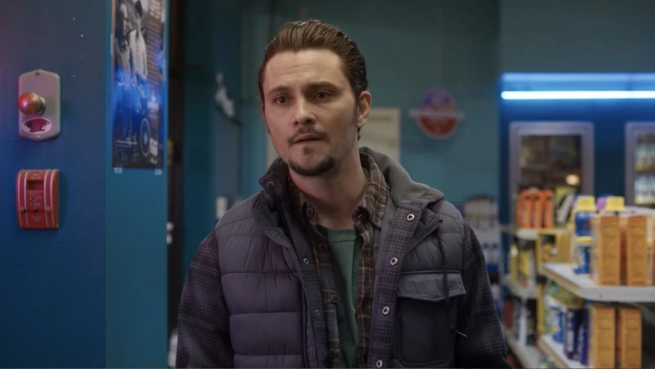 Burn (2019) movie still: medium — A convenience store interior with blue walls, a fire alarm, and blurry stocked shelves. A young man wearing a…