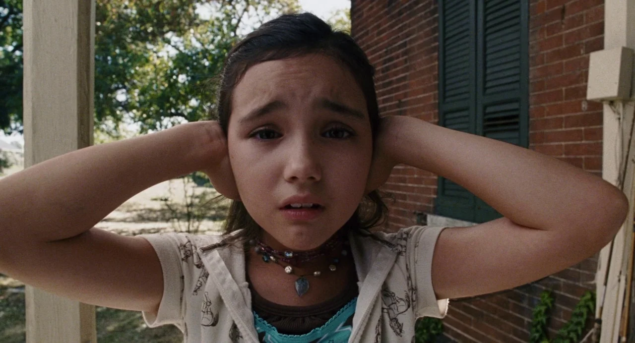 The Happening (2008) movie still: closeup — A young girl stands on a wooden porch in front of a red brick wall. She holds her hands over her ears…