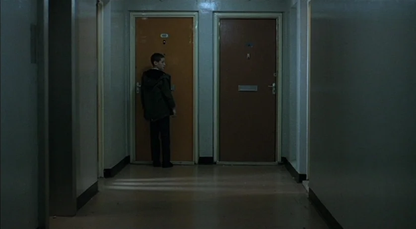 Ratcatcher (1999) movie still: fullbody — A dimly lit apartment hallway features two doors set into pale walls. A young boy stands facing the left…
