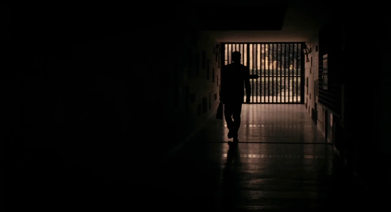 Pasolini (2014) movie still: fullbody — A dark, narrow passageway leads toward a bright outdoor area blocked by a metal gate. A man in a suit walks…