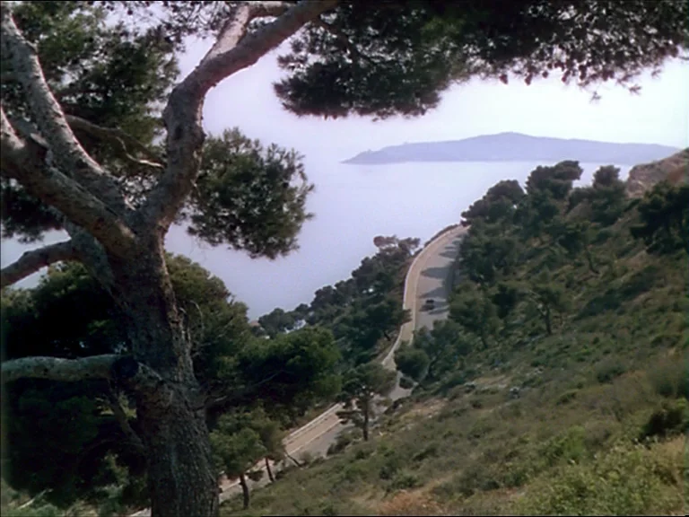 The Red Shoes (1948) movie still: establishing — A winding asphalt road curves along a hillside overlooking a calm sea. Sparse Mediterranean pines…