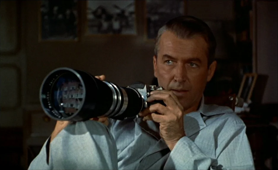 Rear Window (1954) movie still: medium — A dimly lit room decorated with photographs on the wall behind the main subject. A man holds a large…