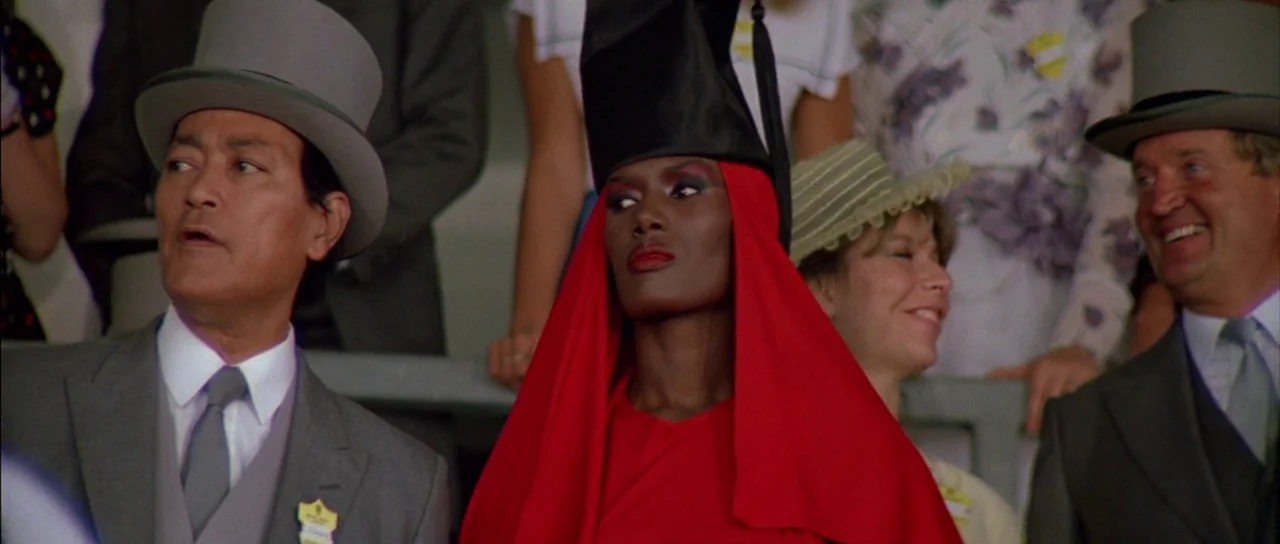A View to a Kill (1985) movie still: medium — A group of people sit in an outdoor grandstand wearing formal attire including top hats. A woman in a…