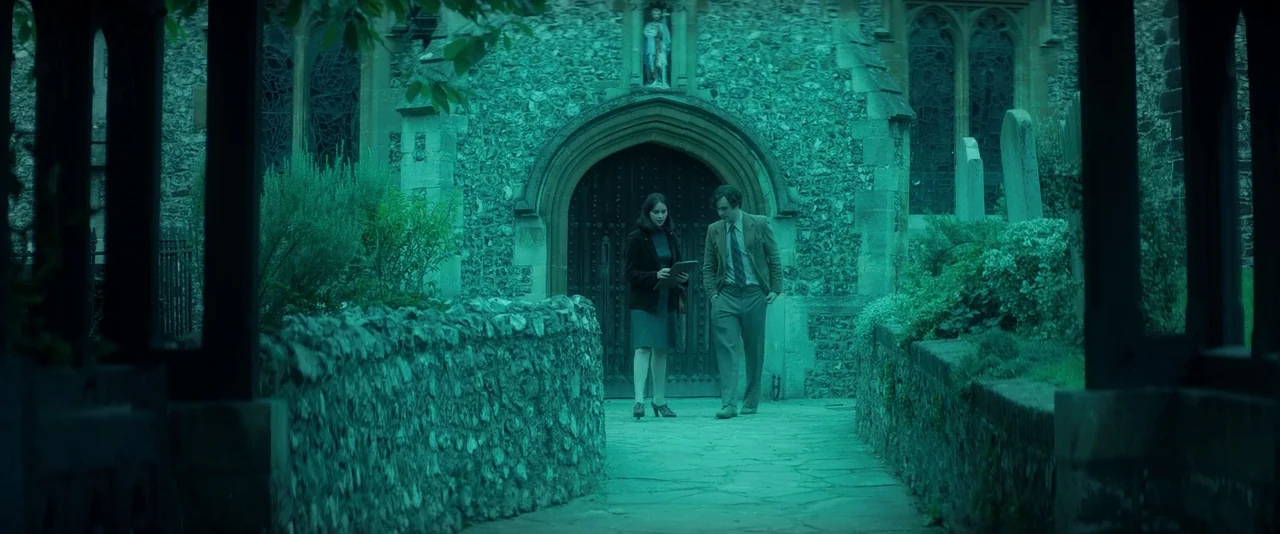The Theory of Everything (2014) movie still: wide — An old stone church with an arched wooden door sits at the end of a path. A man and a woman walk…