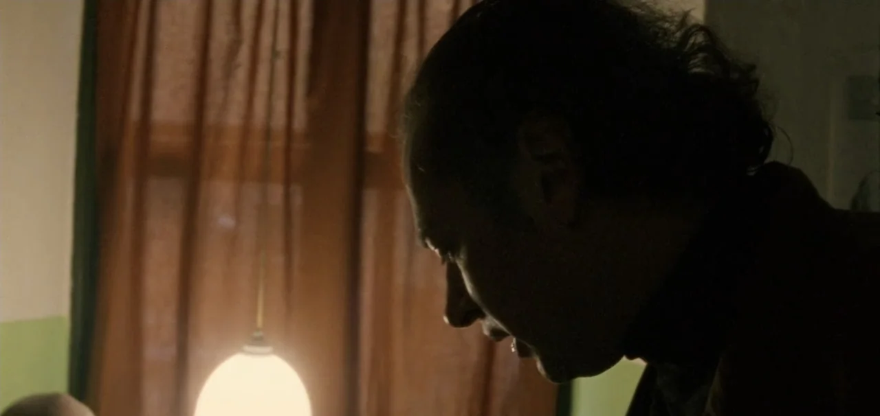 Pusher (1996) movie still: closeup — A dark interior room features a thin curtain over a window and a lamp. A man stands in profile, gazing downward…