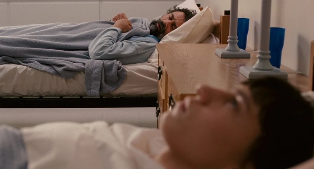 It’s Kind Of A Funny Story (2010) movie still: medium — A minimalist shared bedroom contains two single beds separated by a wooden nightstand. A…