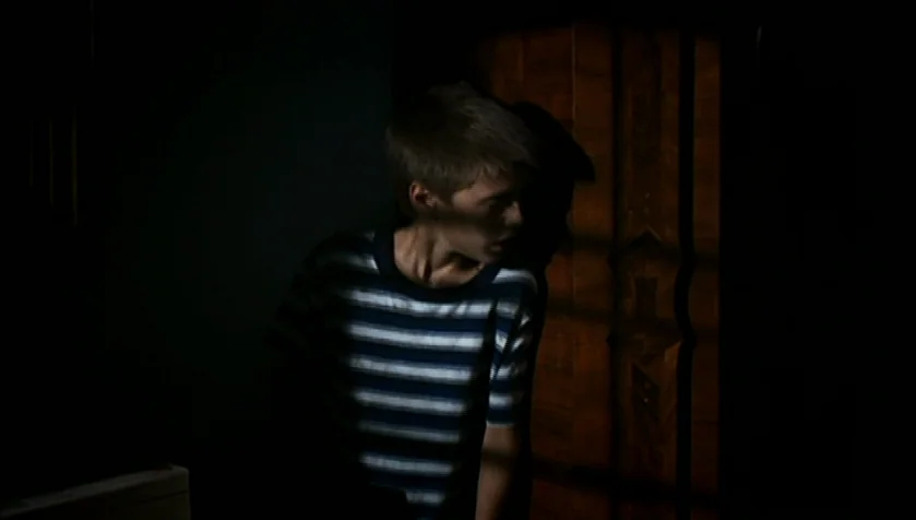 Funny Games (1997) movie still: medium — A dimly lit indoor space features a dark wooden door with vertical paneling detail. A young boy stands…