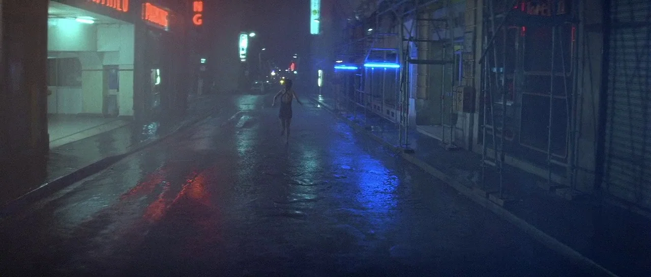 Nikita (1990) movie still: wide — A rain-slicked city street is framed by glowing neon signs and construction scaffolding at night. A lone female…
