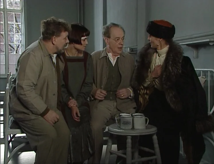 In The Presence of a Clown (1997) movie still: medium — A austere room with pale gray walls and a metal window frame visible. Four individuals are…