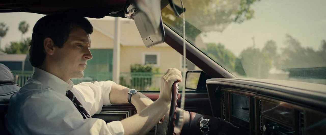 No Man of God (2021) movie still: medium — A man sits inside a vehicle with an aged wooden steering wheel. He drives through a suburban neighborhood…