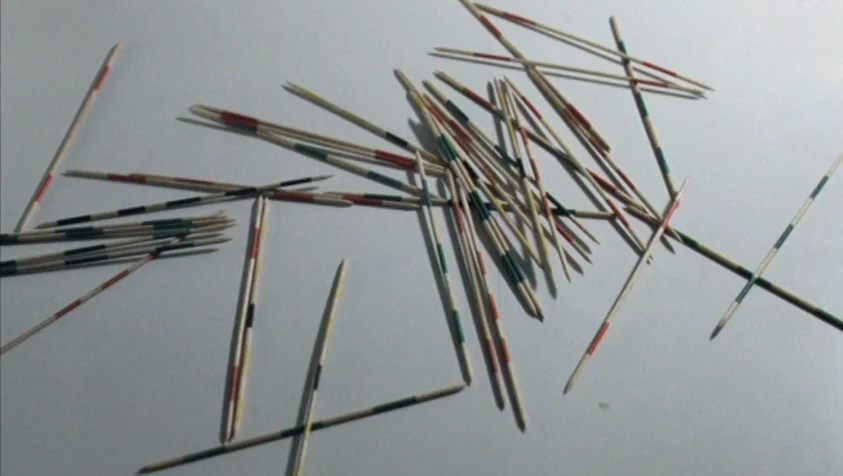 71 Fragments of a Chronology of Chance (1994) movie still: closeup — A pile of multi-colored wooden pickup sticks rests on a smooth, white surface.…