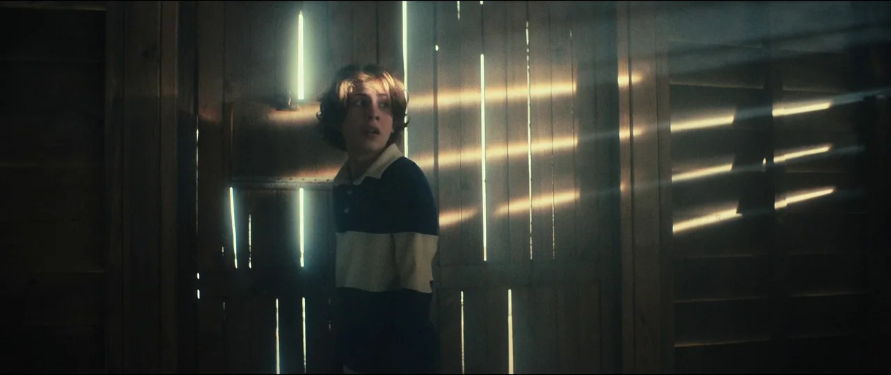 The Toxic Avenger (2023) movie still: medium — A dark wooden shed interior is illuminated by thin horizontal rays of bright sunlight. A teenage boy…