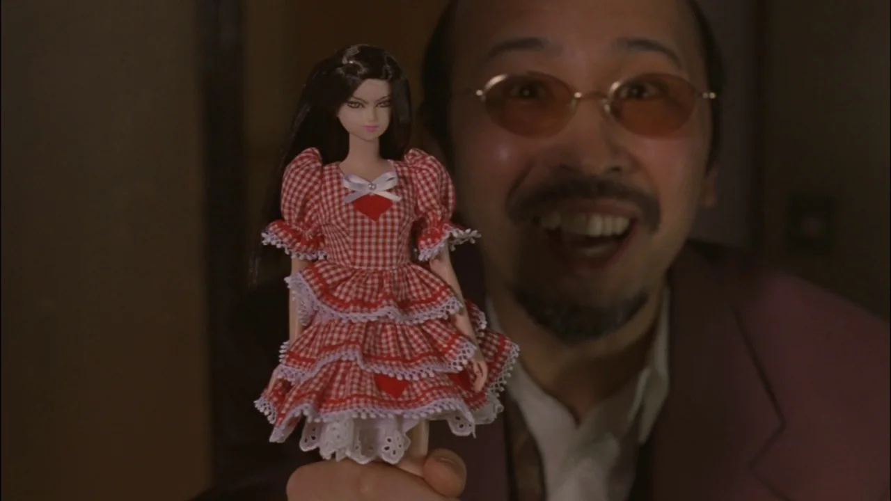 Battle Royale (2000) movie still: closeup — A dimly lit interior space with a man holding a doll in the foreground. The man is positioned out of…