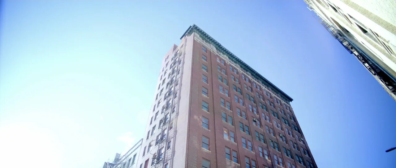 Elevator Game (2023) movie still: establishing — An upward view of a large brick apartment building with a metal fire escape. Clear bright blue sky…
