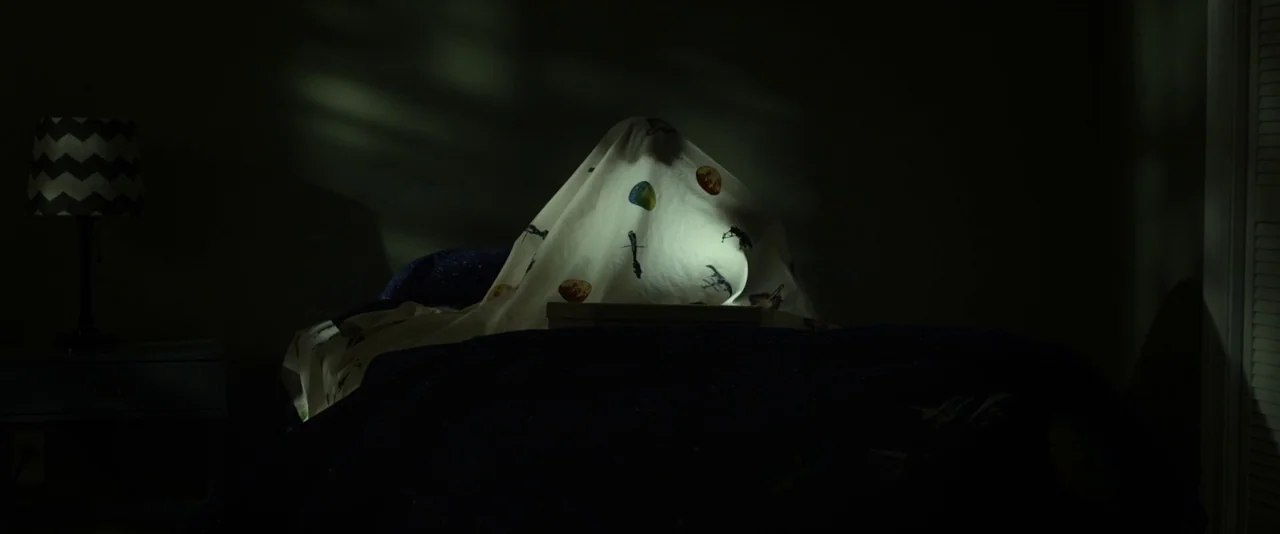 Before I Wake (2016) movie still: medium — A dark, dimly lit bedroom features a bed with a person hiding under a blanket. The blanket is illuminated…
