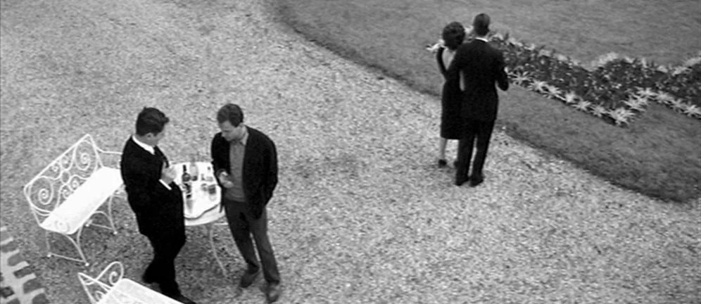 Les Amants (1958) movie still: wide — A gravel-covered courtyard features a small metal table and chairs near a lawn edge. Four people stand in the…