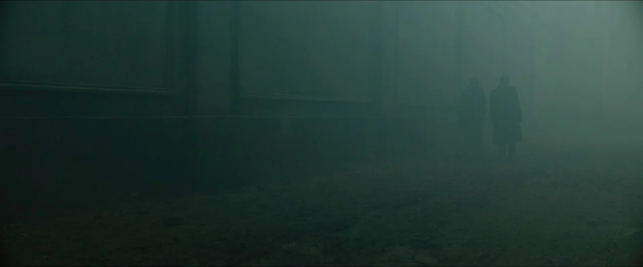 Tchaikovsky’s Wife (2022) movie still: wide — A dark, exterior alleyway is filled with dense, low-hanging fog obscuring the distant walls. Two…