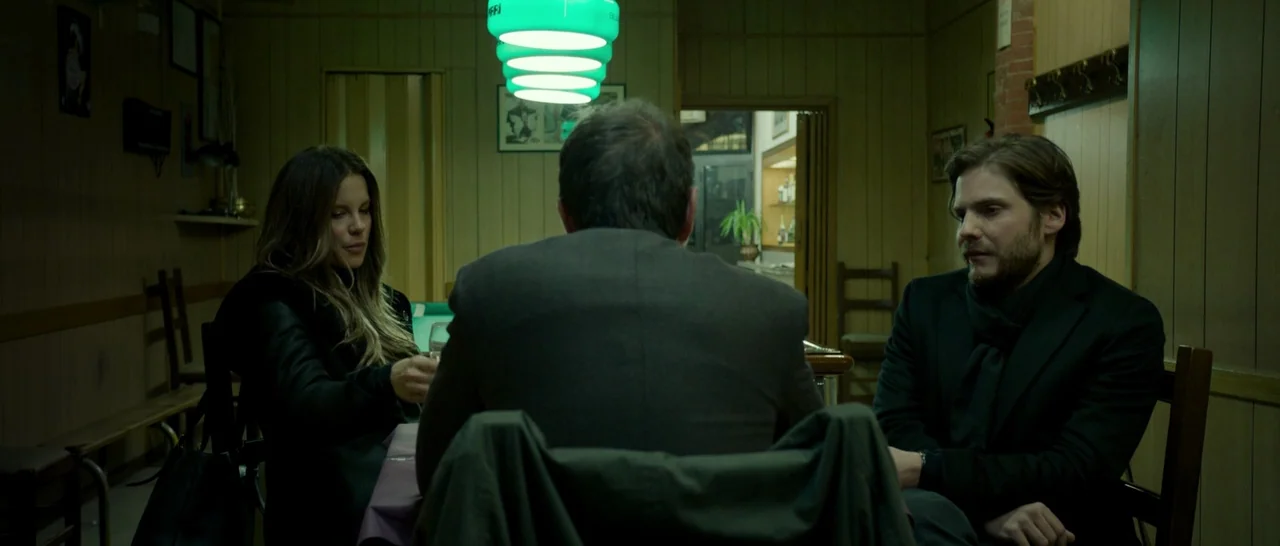 The Face of an Angel (2014) movie still: medium — A dimly lit room with wood-paneled walls and a green hanging light fixture. Two people sit at a…