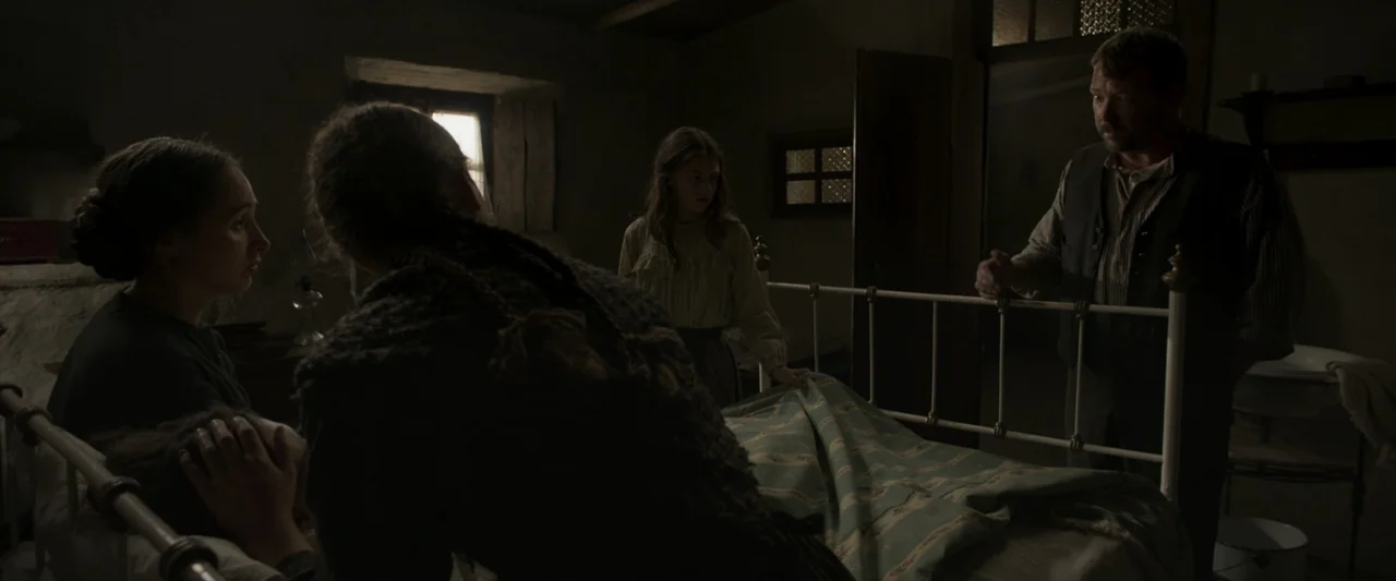 Fatima (2020) movie still: medium — A dark bedroom with bare plaster walls and a simple metal-frame bed. Four people gather around the bed in a…
