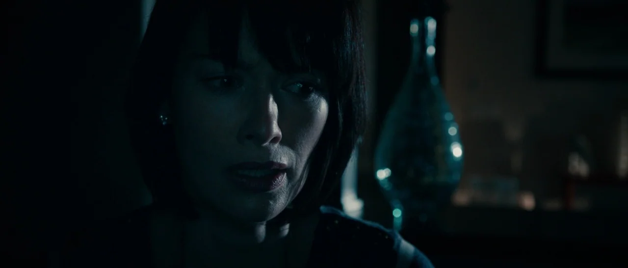 The Purge (2013) movie still: closeup — A dimly lit interior space contains a glass decorative object in the background. A woman looks off-camera…