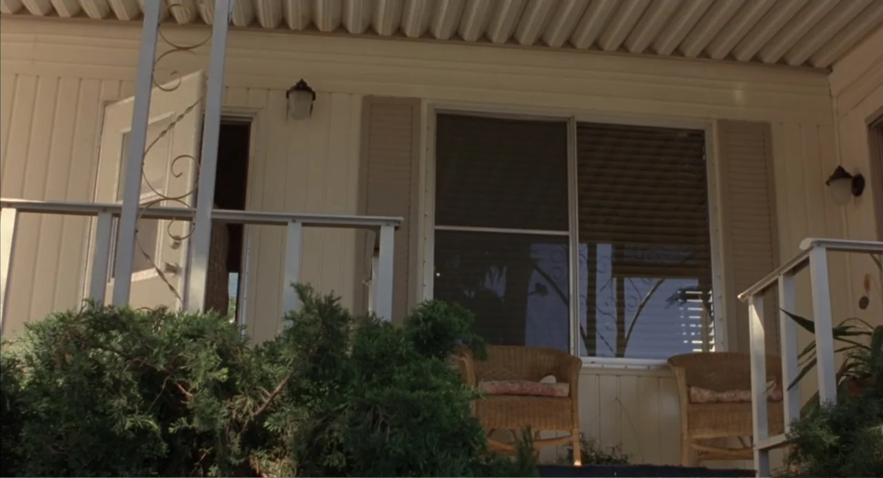 Bogus (1996) movie still: establishing — A cream-colored mobile home features a wooden deck with two wicker chairs. Dense evergreen bushes sit in…