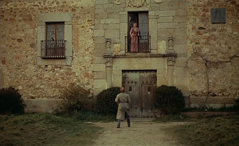 Spirit Of The Beehive (1973) movie still: wide — A stone building with a large wooden door and an upper floor balcony. A man stands in front of the…