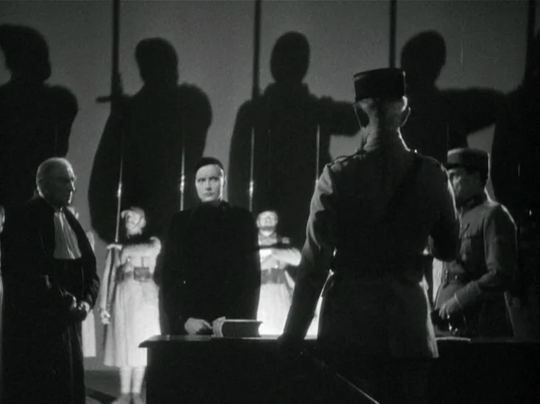 Mata Hari (1931) movie still: medium — A austere courtroom features a woman standing before a desk facing a seated judge. Large, dark silhouettes of…