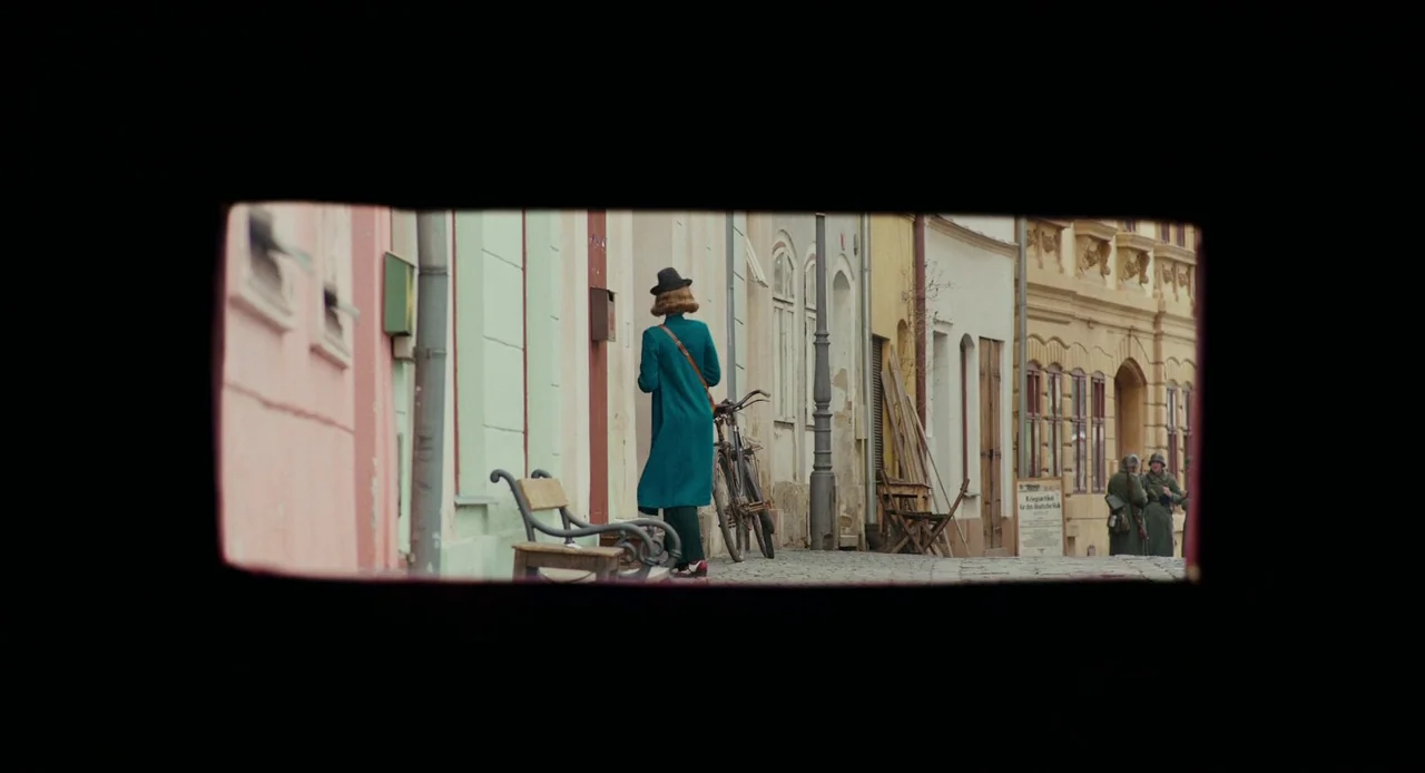 Jojo Rabbit (2019) movie still: wide — A quiet cobblestone street is lined with pastel-colored buildings and a wooden bench. A woman pushes a…