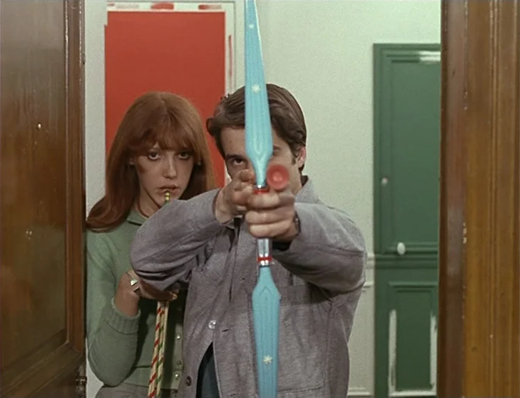 La Chinoise (1967) movie still: medium — A man and woman stand in a brightly lit doorway between rooms with colorful doors. The man holds a plastic…