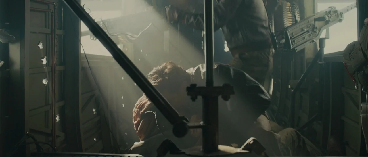 Unbroken (2014) movie still: medium — The interior of a military aircraft shows visible bullet holes in the metal walls. One man remains hunched…