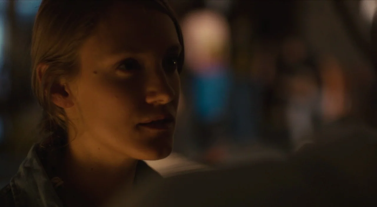 The Comedy (2012) movie still: closeup — A dark indoor space with blurred shapes visible in the out-of-focus background. A young woman's face is lit…
