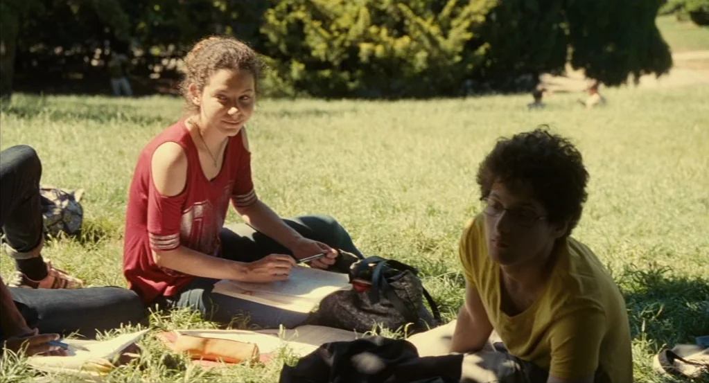 Things to Come (2016) movie still: medium — Two students sit on the grass in a sunny park surrounded by leafy trees. The woman writes in a notebook…