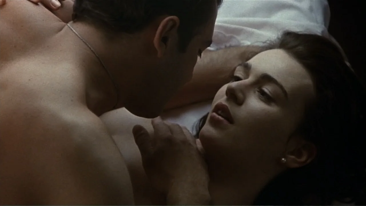 The Mambo Kings (1992) movie still: closeup — A dark bedroom interior shows two people lying on white sheets. The man faces the woman while she…