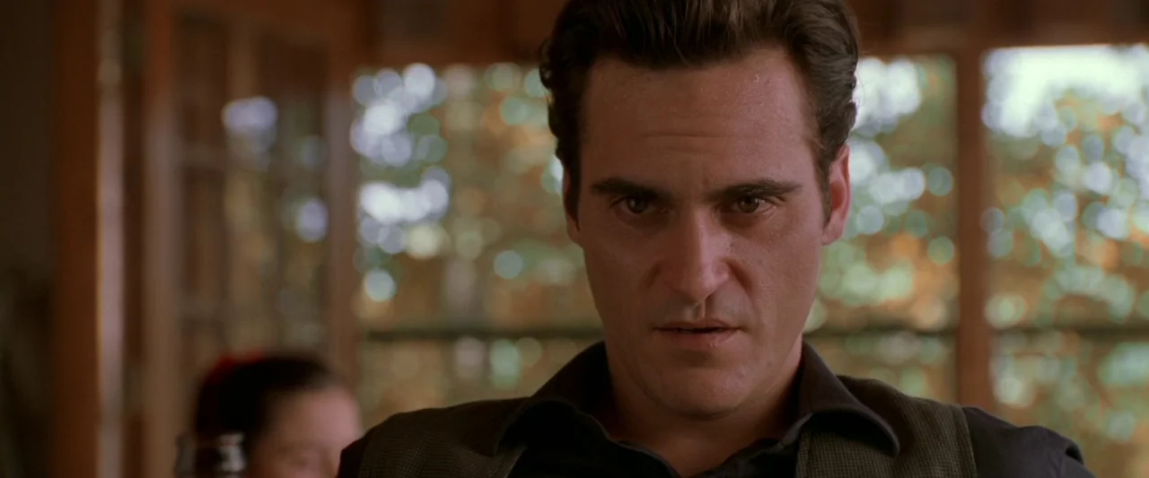 Walk The Line (2005) movie still: closeup — A man stands inside a house with a large window and blurred foliage behind him. He looks toward the…