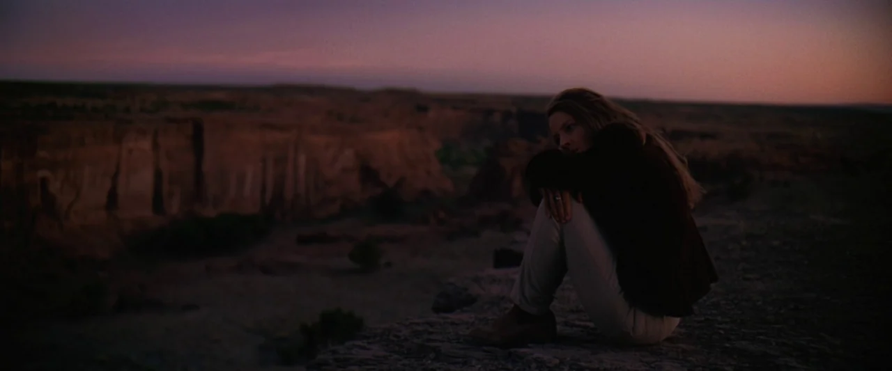 Contact (1997) movie still: wide — A woman sits at the edge of a rocky canyon during the dusk. She sits with her knees pulled to her chest looking…