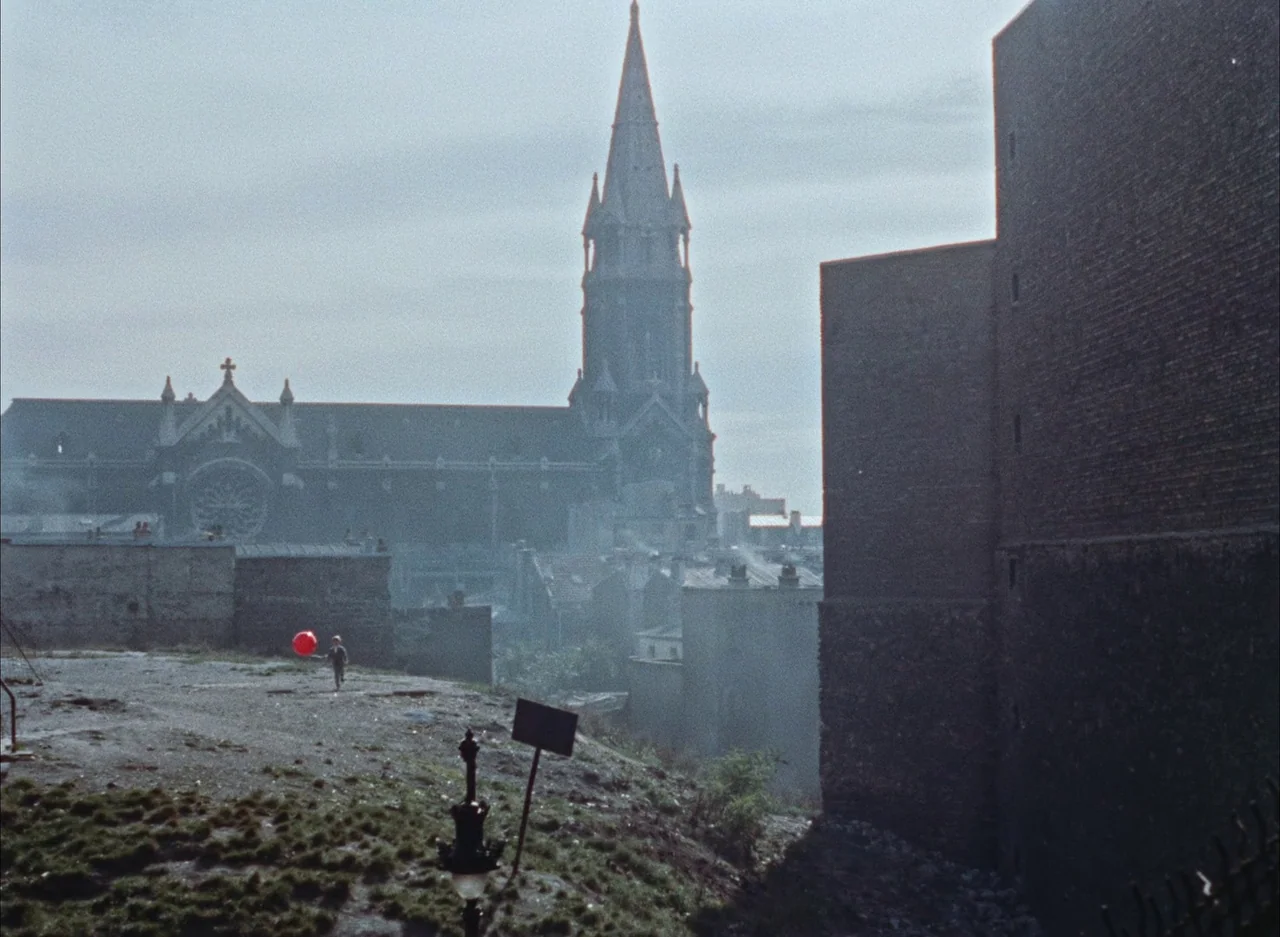 The Red Balloon (1956) movie still: wide — An urban hilltop lot looks down over a hazy cityscape featuring a large stone church. A small boy holding…