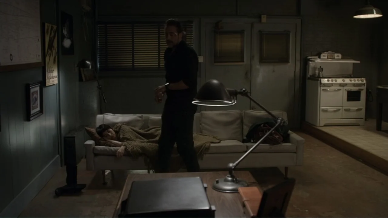The Messenger (2015) movie still: medium — A dimly lit room features a white sofa, a vintage stove, and dark walls. A man in dark clothing stands…