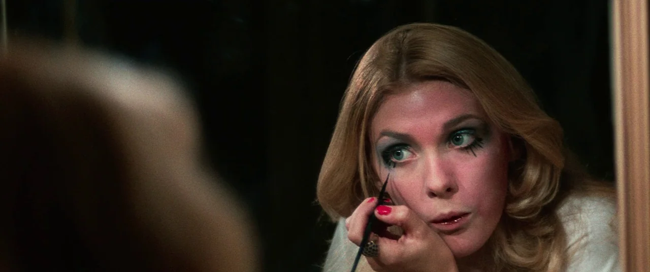 Death Game (1977) movie still: closeup — A woman stares into a mirror while applying dramatic black makeup around her eyes. She holds a small…