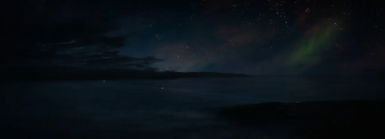 28 Years Later (2025) movie still: establishing — A dark coastline stretches across the frame at night beneath a star-filled sky. Faint aurora…