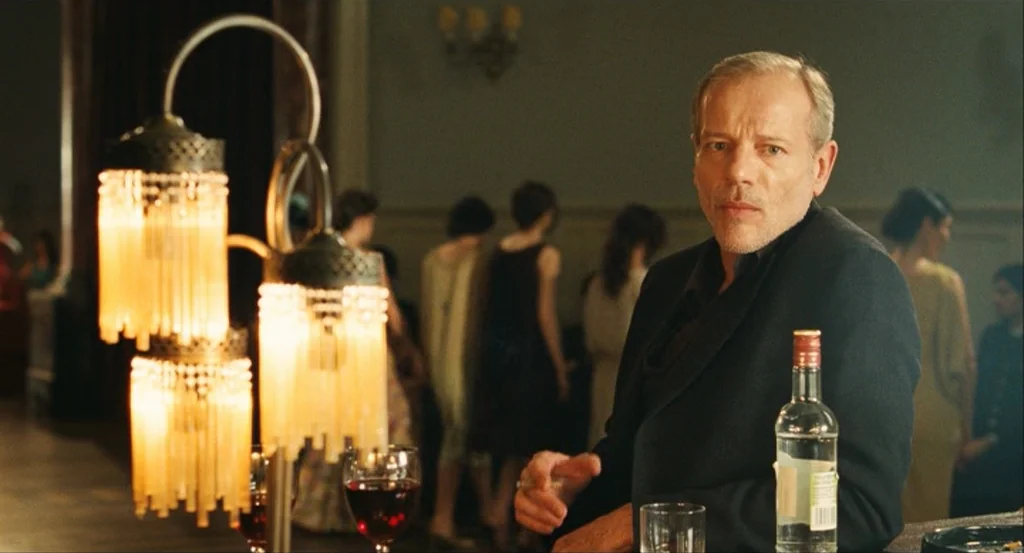 This Night (2008) movie still: medium — A dimly lit bar contains patrons in formal wear standing in the blurred background. A middle-aged man sits…
