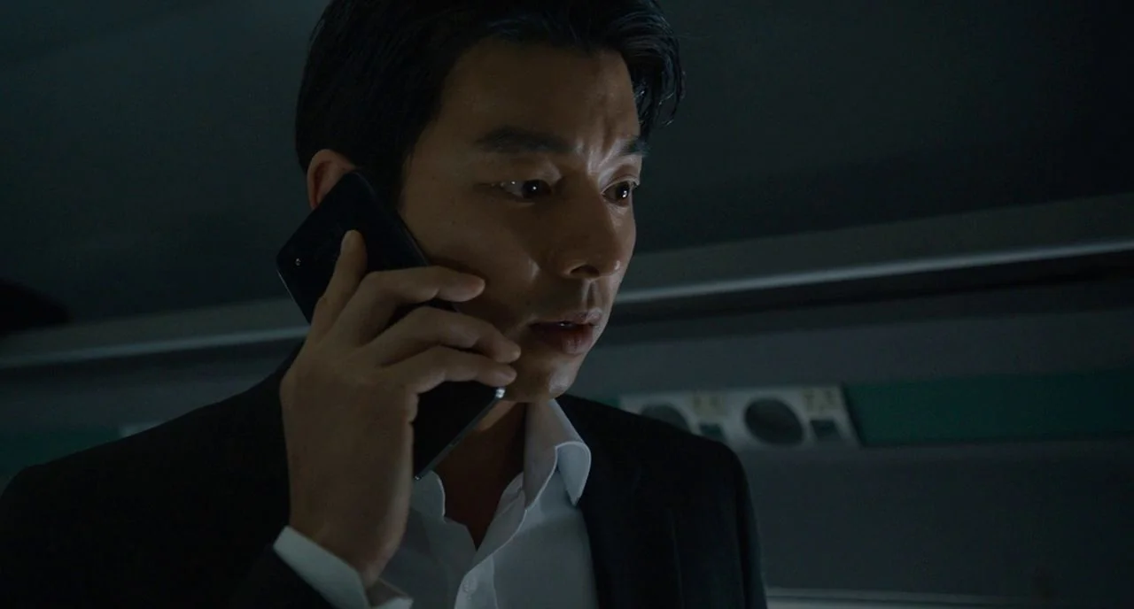 Train To Busan movie still: closeup — The interior of a dark train car with indistinct features in the background. A man in a suit listens to a…