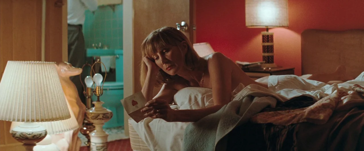 The Adjuster (1991) movie still: medium — A dimly lit hotel room features red walls and a glimpse of a teal bathroom. A woman lies on the bed…