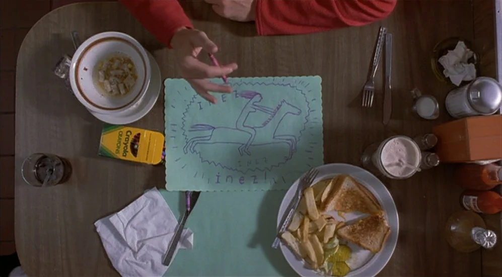Bottle Rocket (1996) movie still: wide — A wooden table is set with a half-eaten meal, cutlery, and a crayon drawing. A person's hand reaches into…