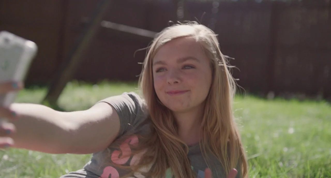 Eighth Grade (2018) movie still: medium — A teenage girl sits on a grassy lawn during the day. She extends her arm to take a selfie with a smartphone.