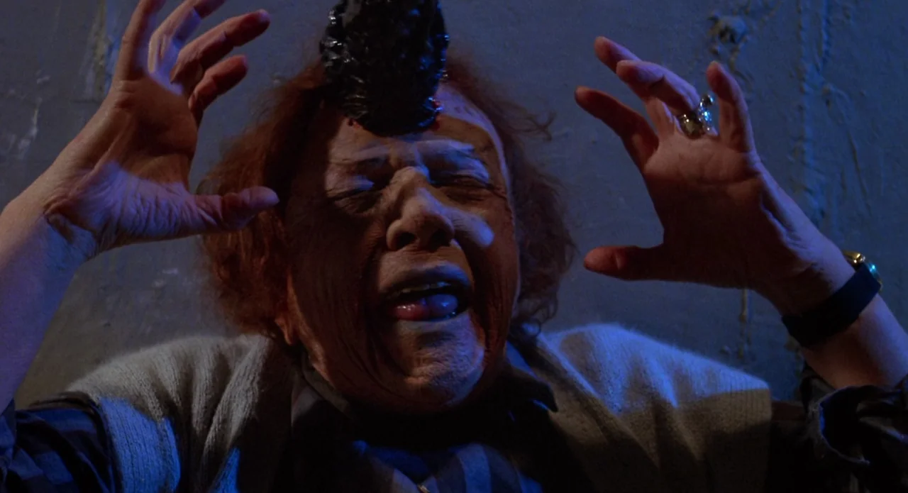 Brain Damage (1988) movie still: closeup — An interior space with a plain dark wall illuminated by a cool blue light. An elderly person exhibits a…