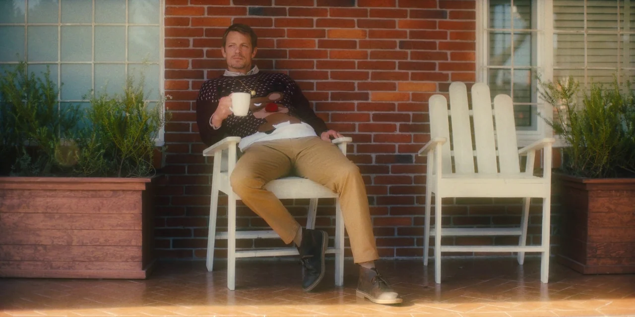 Silent Night (2023) movie still: medium — A man sits on a white wooden chair on a paved porch with a brick wall backdrop. He holds a white coffee…