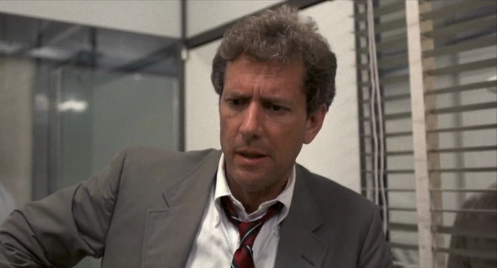 Salvador (1986) movie still: medium — A modern 1980s workspace features horizontal window blinds and paneled walls. A middle-aged man in a suit…