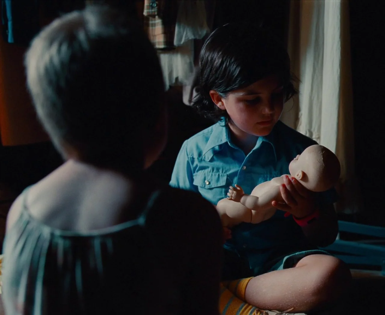 Monica (2022) movie still: medium — An interior space with a dimly lit background featuring draped fabrics and clothing items. A young boy holds a…