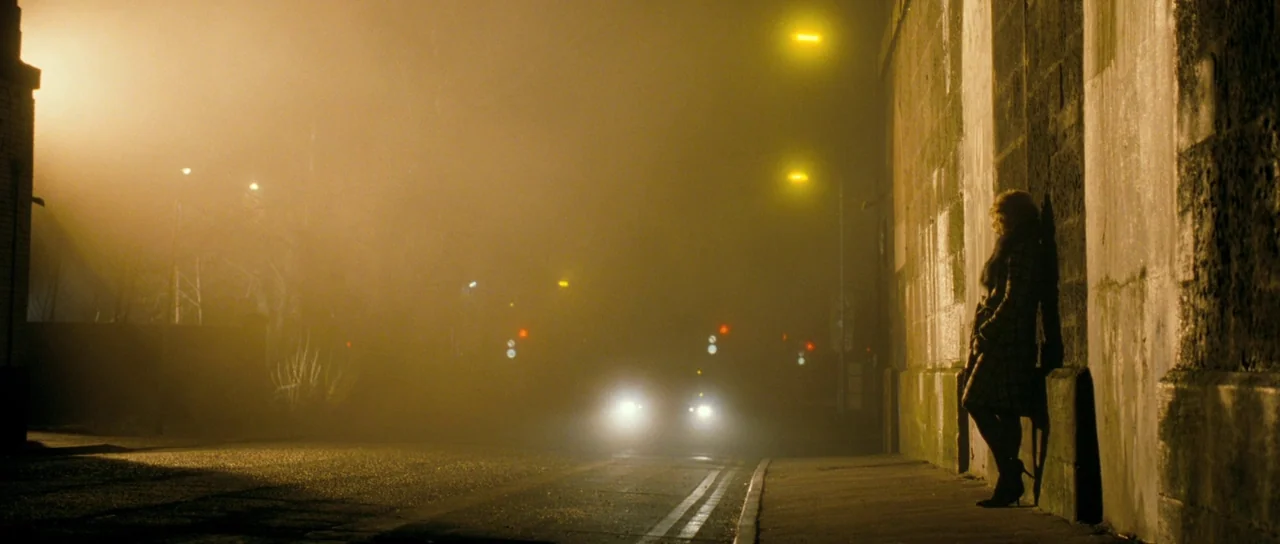 Filth (2013) movie still: wide — A thick, hazy fog obscures a nocturnal road passing beneath a massive stone bridge. A woman stands stationary…
